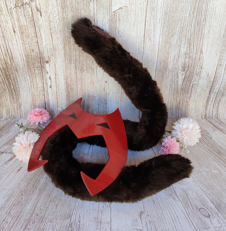 May include: A brown faux fur tail with a red and black plastic mask attached. The mask has a sharp, angular design with a pointed top and a wide, open mouth.