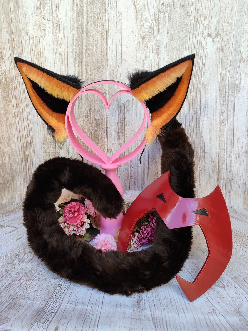 May include: A brown faux fur tail, a pair of orange and black cat ears, and a red mask with black accents. The cat ears are on a headband and the tail is curled up. The mask is shaped like a crescent moon.