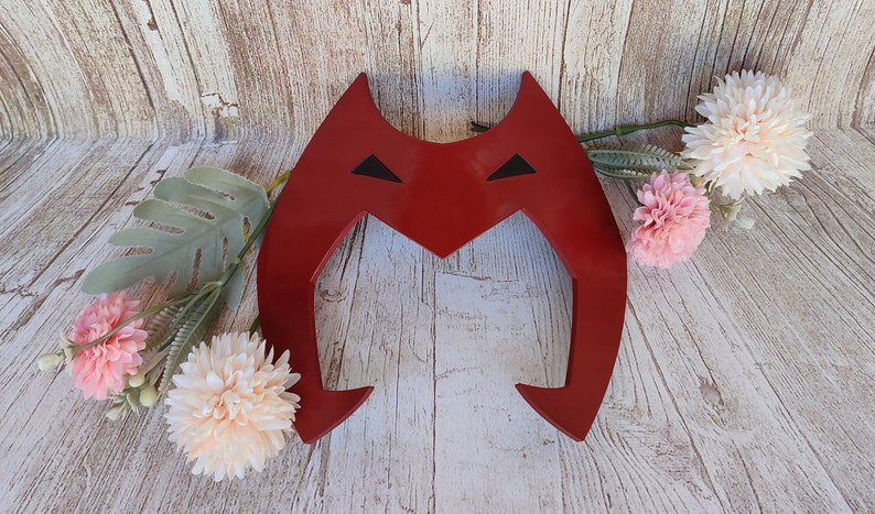 May include: Red plastic headband with a stylized M shape and black eyes. The headband is on a wooden background.