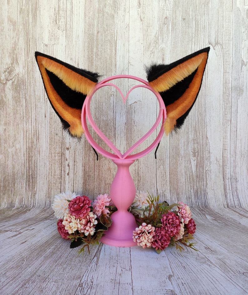 May include: A pair of black and orange faux fur cat ears on a pink heart-shaped headband stand. The stand is on a wooden surface with pink flowers.