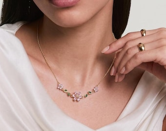Pode incluir: Um colar de corrente em tom dourado com um motivo floral central de pétalas rosa claro e pequenas folhas verdes. O colar é usado ao redor do pescoço, exibindo um design delicado com três elementos florais distintos conectados por hastes douradas finas.