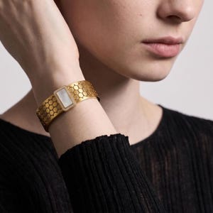 May include: A gold-toned cuff bracelet featuring a textured honeycomb pattern across the band. The centre of the bracelet displays a rectangular white mother-of-pearl inlay framed by a border of small, clear, sparkling stones set in gold.