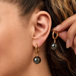 May include: A pair of gold hoop earrings featuring a dangling dark gray spherical pearl. One earring is attached to the earlobe, while the other is held up to show the circular gold hoop design and the smooth, reflective surface of the dark pearl.