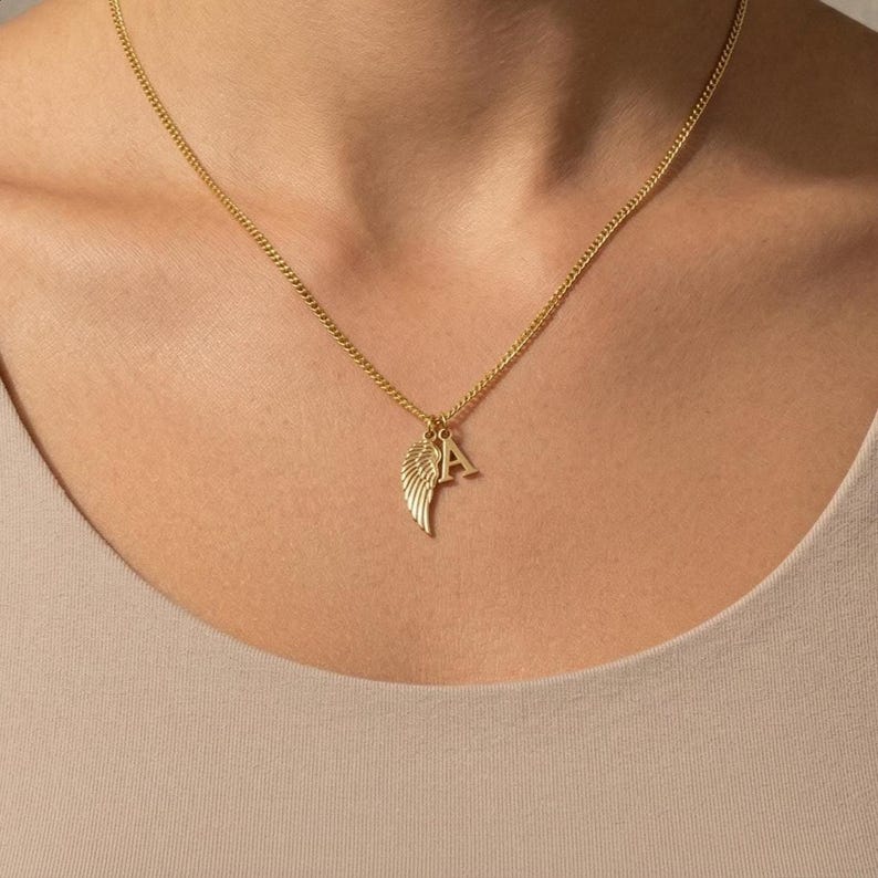May include: Gold-coloured necklace with a delicate chain. The pendant features a wing and the letter "A". The necklace is worn around the neck.