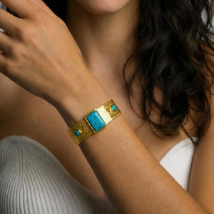 May include: A gold-toned cuff bracelet featuring a textured surface with a large rectangular turquoise-colored stone in the center and two smaller round turquoise-colored stones on either side, set within a wide, ornate metal band.
