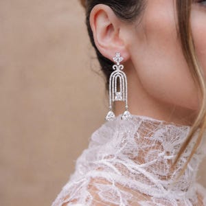 May include: A silver-toned dangle earring featuring a vertical arch design encrusted with small clear stones. The earring includes a central rectangular stone and two dangling triangular stones at the base, all set in a metallic frame.