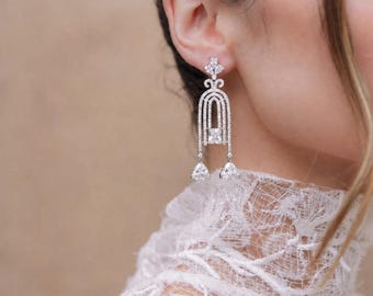 May include: A silver-toned dangle earring featuring a vertical arch design encrusted with small clear stones. The earring includes a central rectangular stone and two dangling triangular stones at the base, all set in a metallic frame.