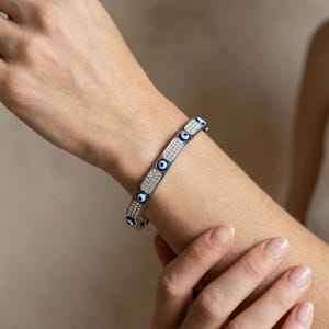 May include: Silver-toned bangle bracelet featuring alternating blue evil eye charms and sections of sparkling white crystals. The bracelet is shown on a person's arm, showcasing its design and details. A stylish piece of jewelry.