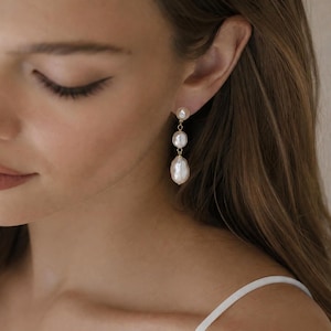 May include: A drop earring featuring three white baroque pearls of varying sizes arranged vertically. The pearls are connected by thin gold-toned metal links, with the smallest pearl at the top and the largest, elongated pearl at the bottom.