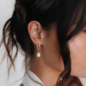 May include: A gold-toned drop earring featuring two interlocking oval links, with the upper link accented by small clear stones. A single white teardrop-shaped pearl hangs from the bottom link. A small stud earring is visible in the upper ear lobe.