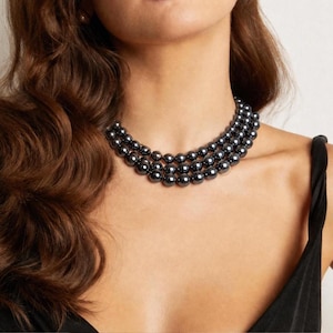 May include: A triple-strand necklace featuring uniform, round, dark grey metallic beads with a high-gloss finish. The beads are strung together in three parallel rows, creating a layered, collar-style appearance that rests against the base of the neck.