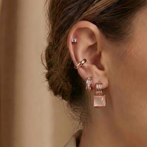 May include: An ear featuring multiple pieces of jewellery. A small square stud, a curved ear cuff, and a pair of hoop earrings with a square, light pink stone are visible. The jewellery is rose gold in colour.
