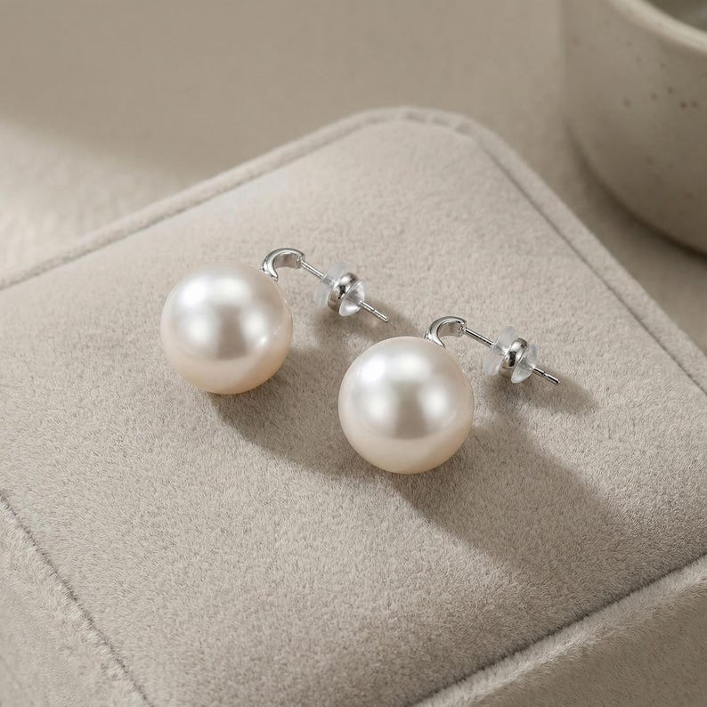 May include: A pair of stud earrings featuring large, round, lustrous white pearls. Each pearl is securely mounted on a polished silver-tone metal post with a clear plastic backing, designed for pierced ears with a simple, clean aesthetic.