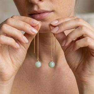 Peut inclure: Une paire de boucles d'oreilles pendantes dorées composées de fines chaînes retenant des perles rondes et lisses de couleur vert pâle. Chaque chaîne est surmontée d'une tige métallique droite conçue pour être glissée à travers le lobe de l'oreille.