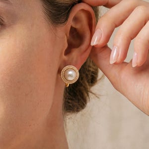 Peut inclure: Une boucle d'oreille à clip dorée présentant une perle blanche ronde centrale entourée d'une bordure circulaire texturée avec un contour perlé. La boucle d'oreille est fixée au lobe par un mécanisme à clip métallique visible à l'arrière.