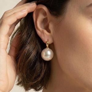 May include: A single gold hoop earring featuring a large, round, white pearl. The earring is attached to the ear. The pearl is a creamy white colour and has a smooth, polished surface. The gold hoop is small and simple.