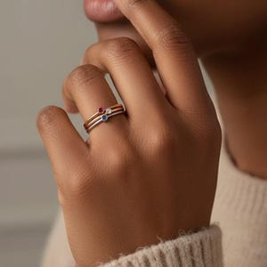 May include: Three delicate rings stacked on a finger. The top ring is gold with a small red gemstone, the middle ring is gold, and the bottom ring is silver with a small blue gemstone. The rings are simple and elegant.