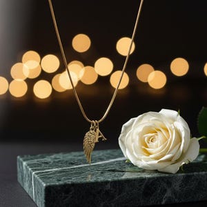 May include: A gold necklace with a pendant shaped like an angel wing and the letter "A". The necklace is presented on a green marble surface, next to a white rose. The background features blurred golden lights.