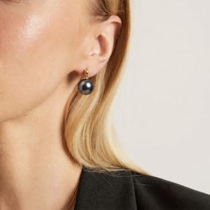 May include: A single drop earring featuring a smooth, spherical dark gray pearl suspended from a polished gold-tone metal hook. The earring is worn on the earlobe, showcasing the contrast between the metallic gold setting and the dark pearl.