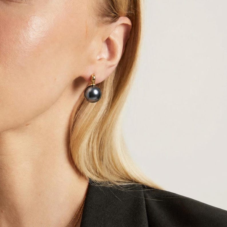 May include: A single drop earring featuring a smooth, spherical dark gray pearl suspended from a polished gold-tone metal hook. The earring is worn on the earlobe, showcasing the contrast between the metallic gold setting and the dark pearl.