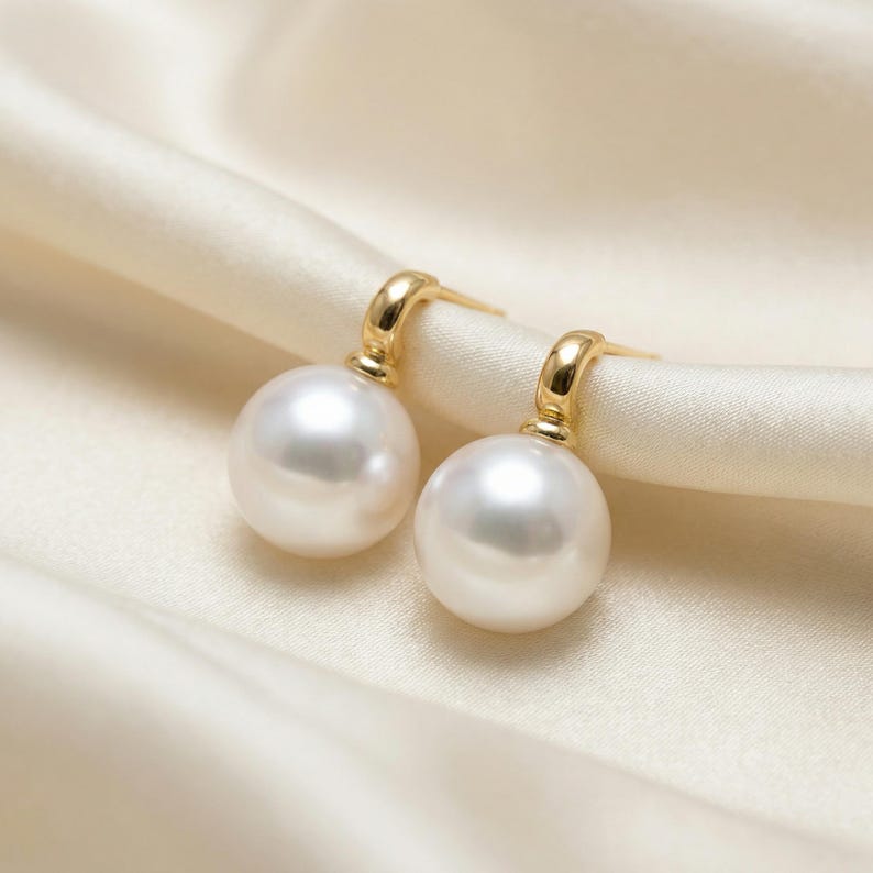 May include: A pair of drop earrings featuring two round, white, lustrous pearls suspended from polished gold-tone metal hooks. The earrings have a smooth, reflective surface and a classic design, showcasing the spherical shape of each pearl.