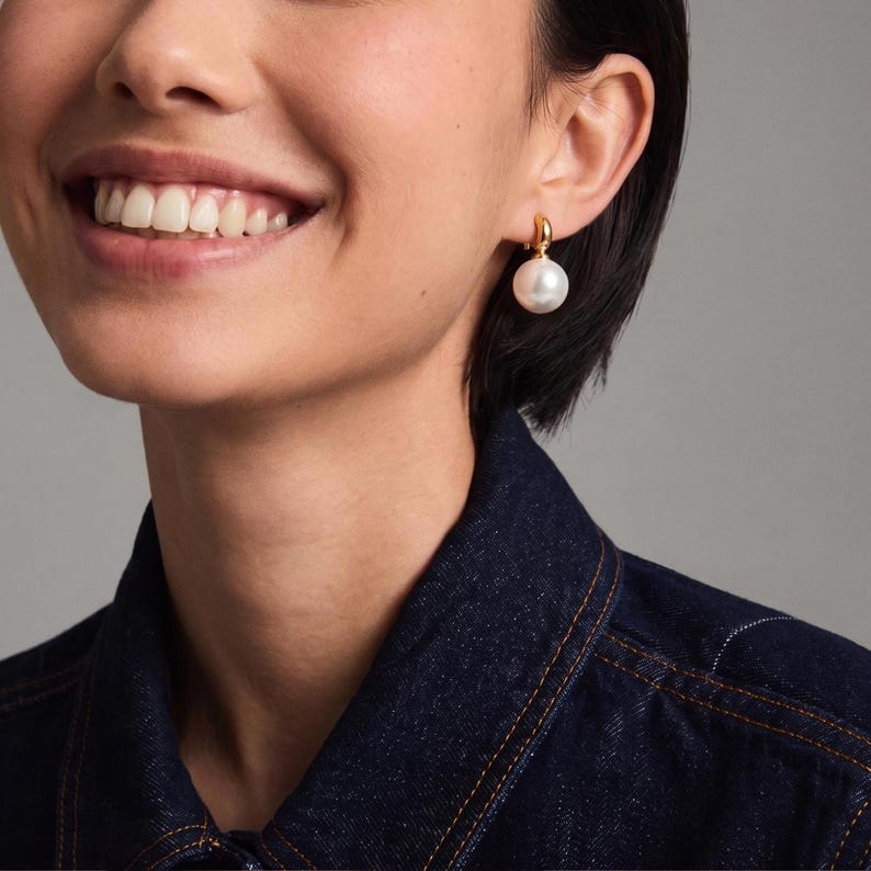 May include: A gold hoop earring featuring a single round white pearl drop. The earring is secured with a small gold clasp that connects the hoop to the pearl, creating a simple and elegant drop design that hangs from the earlobe.