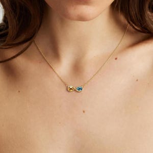 May include: A gold chain necklace featuring an infinity-shaped pendant. The pendant is set with two pear-shaped gemstones, one yellow citrine and one blue topaz, connected in a continuous loop design that rests against the collarbone area.