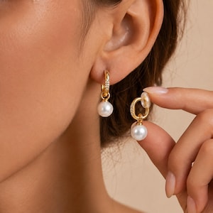 May include: A pair of gold hoop earrings featuring a small, round white pearl drop. The gold hoops are accented with tiny clear stones along the front edge. One earring is worn on the earlobe, while the other is held to show the design.