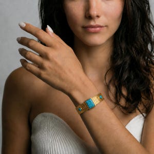 Op de afbeelding: Een goudkleurige armband met een centrale rechthoekige turkoois steen en twee kleinere ronde turkoois stenen aan weerszijden. Het metalen oppervlak van de armband heeft een ingewikkelde, reli&euml;ftextuur over de gehele lengte.