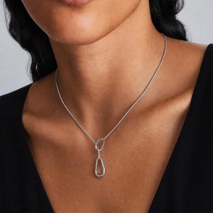 May include: A silver-toned necklace featuring a thin, flexible chain and a pendant composed of two interconnected, irregular, organic-shaped loops. The metal has a polished, reflective finish, and the design hangs centrally at the collarbone.