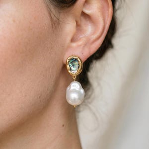 Op de afbeelding: Gouden oorhangers. Elke oorbel heeft een goudomrand, ovaal abalone schelp accent aan de bovenkant en een grote, witte barokparel die eronder hangt. De oorbellen zijn ontworpen om op het oor te worden gedragen.
