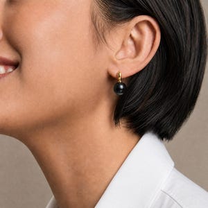 May include: A gold hoop earring featuring a single, smooth, spherical black bead dangling from the bottom. The earring is secured to the earlobe with a small gold clasp, creating a simple and polished look with a high-contrast color palette.