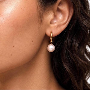 May include: A gold hoop earring featuring a single, smooth, round, pale pink pearl drop. The earring is secured through the earlobe, with the polished gold hoop connecting to the pearl via a small, circular gold link, creating a simple drop design.