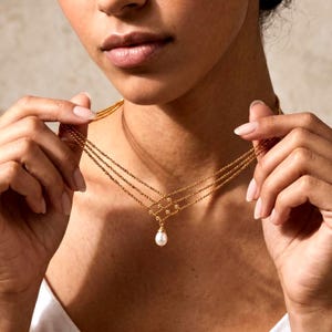 May include: A gold-toned multi-strand necklace featuring three delicate rope chains that converge into a central decorative lattice pattern. A single, small, white teardrop-shaped pearl pendant hangs from the center of the gold metal structure.