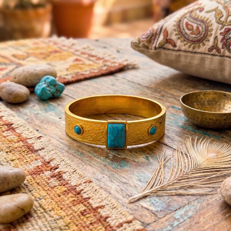 Op de afbeelding: Een goudkleurige metalen armband met een ingewikkeld ge&euml;tst patroon over de band. De armband is geaccentueerd met drie rechthoekige en ronde turkooiskleurige stenen die met regelmatige tussenpozen in het metalen oppervlak zijn gezet.