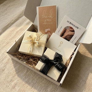 May include: A white cardboard shipping box containing two square gift boxes, one with a cream ribbon and one with a black ribbon, surrounded by paper filler. Includes a brown thank you card, an Auroglow jewelry studio card, and a white card with a tulip icon.