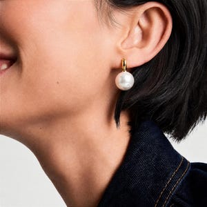 Op de afbeelding: Een gouden oorring met een enkele, grote, ronde witte parel als hanger. De oorbel is bevestigd met een kleine goudkleurige metalen lus die de parel met de oorlel verbindt, wat zorgt voor een klassiek ontwerp van een hangende oorbel.