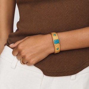 Pode incluir: Uma pulseira de punho em tom dourado com uma pedra turquesa retangular central e duas pedras turquesas circulares menores incrustadas numa banda de metal texturizada, acompanhada por um anel de banda dourado simples no dedo.