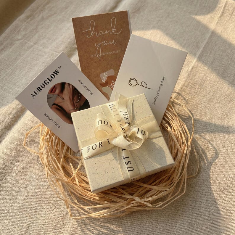 Op de afbeelding: Een vierkante, gespikkelde gebroken witte geschenkdoos met een lint waarop de tekst JUST FOR YOU staat, genesteld in lichtbruin raffia. Inbegrepen zijn een bruine bedankkaart, een witte kaart met sieradenpoetsdoek en een AUROGLOW-sieradenkaart.