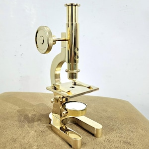 Può includere: Un microscopio in ottone dorato con una manopola rotonda sul lato e un palco circolare. Il microscopio è su una superficie marrone.