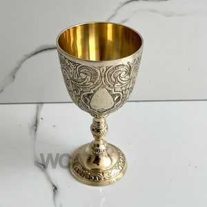 Może przedstawiać: Złoty metalowy kielich z ozdobnym wzorem. Kielich ma okrągłą podstawę i wysoką, smukłą nóżkę.