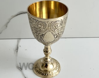 May include: A gold-colored metal goblet with an ornate design. The goblet has a round base and a tall, slender stem.