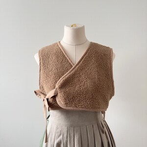 May include: A brown, sleeveless wrap vest made of a textured, fuzzy material. The vest has a V-neck and is trimmed with a smooth, light brown fabric. It ties at the side with matching fabric ties. The vest is displayed on a mannequin.