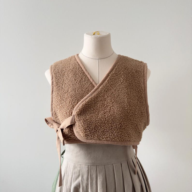 May include: A brown, sleeveless wrap vest made of a textured, fuzzy material. The vest has a V-neck and is trimmed with a smooth, light brown fabric. It ties at the side with matching fabric ties. The vest is displayed on a mannequin.