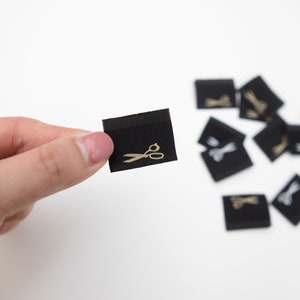 May include: Black fabric label with a gold scissor icon. The label is being held by a hand.