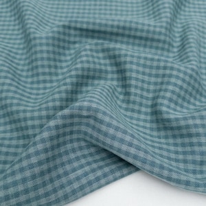 May include: Close-up of a teal and white checked fabric. The material has a soft, textured appearance with a classic gingham pattern. The fabric is folded, showing the weave and colour variations.