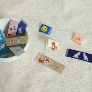 May include: An assortment of colourful fabric labels with various embroidered designs, including a smiling face, a penguin, a fox, a cat, a dinosaur, a dog, and a cherry. The labels are in a bowl and on a beige surface. One label reads "Petit, Petite".