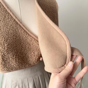 May include: A tan-colored vest with a soft, textured exterior and a smooth, light brown interior. The vest has a wrap-around design with a tie closure. The garment is displayed on a mannequin.
