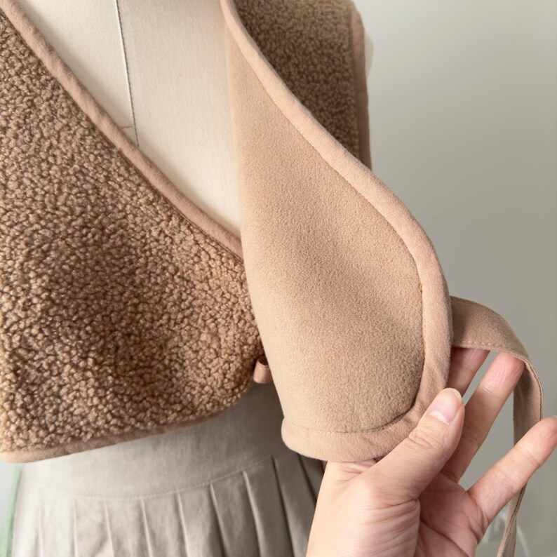 May include: A tan-colored vest with a soft, textured exterior and a smooth, light brown interior. The vest has a wrap-around design with a tie closure. The garment is displayed on a mannequin.