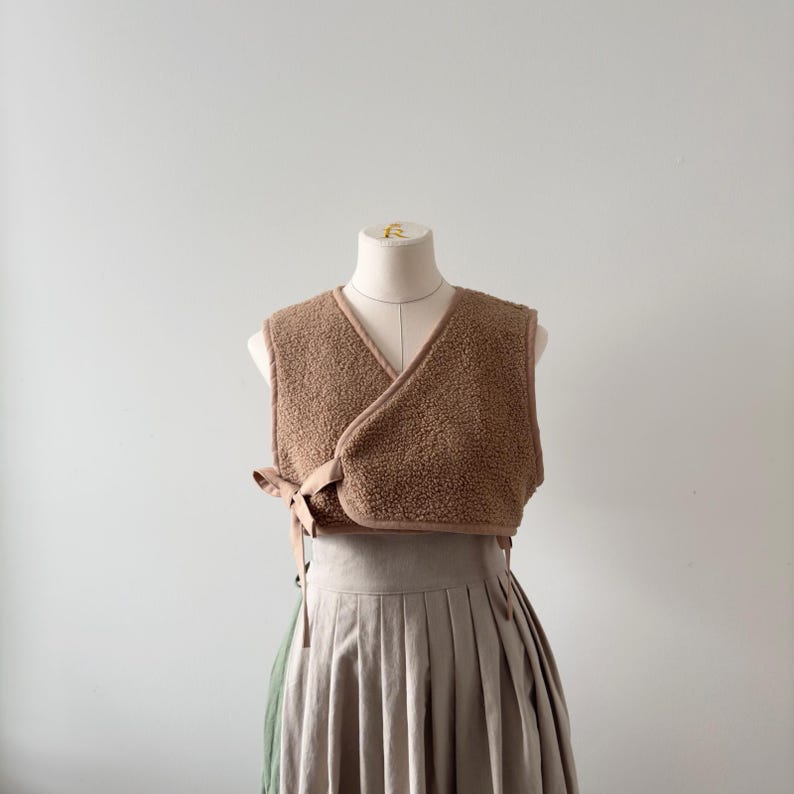 May include: A brown, sleeveless wrap vest made of a soft, textured material. The vest has a V-neck design and ties at the waist with a matching fabric. The vest is displayed on a mannequin, paired with a pleated skirt.
